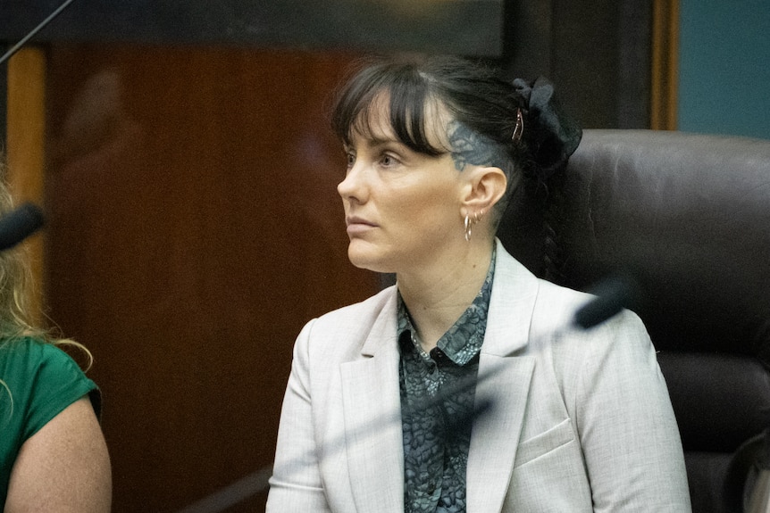 Kat McNamara, white person, long-hair tied back in bun, with gray shirt and pink blazer sitting in brown leather seat.