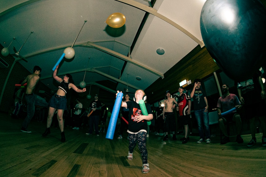 A young child wearing ear muffs looks up at a yellow balloon as people dance around them.