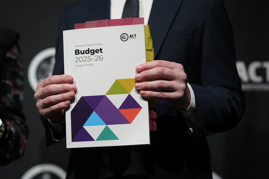 An unidentifiable man's hands hold an A4 book with a title reading "Australian Caption Territory Budget 2025-26".