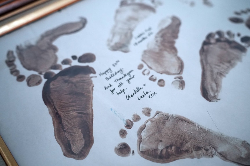 A child's footprints in brown paint on white paper in a gold frame.