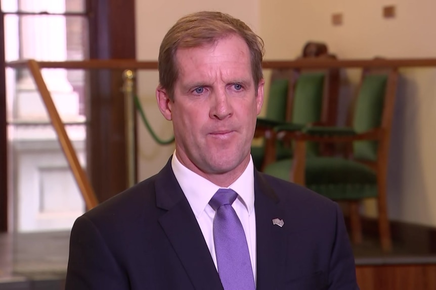 A man in a black suit, pink shirt and purple tie stands looking seriously at the camera