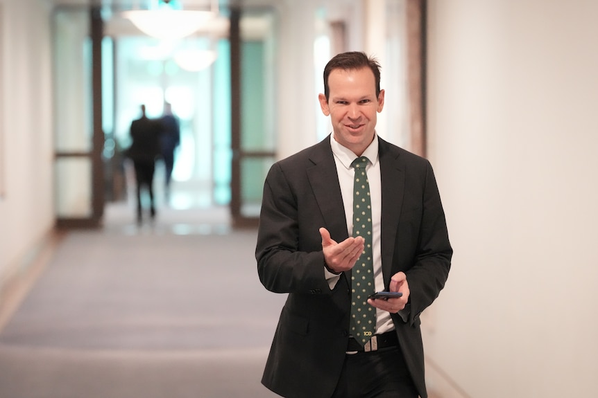 A dark-haired man in a dark suit walks down a corridor.