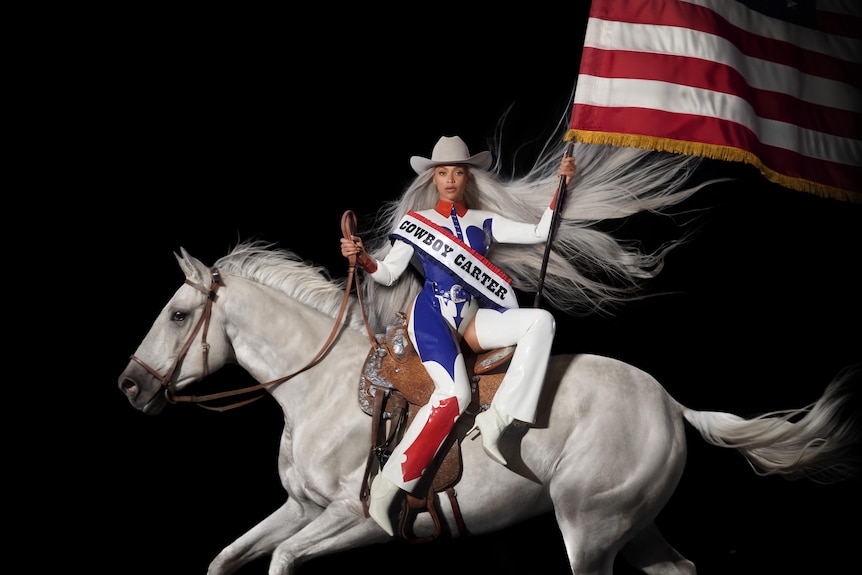 Beyonce on a white horse, carrying an American flag and wearing red, blue and white, with a white cowboy hat on