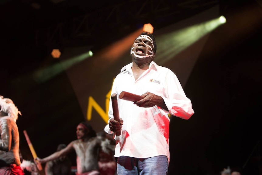 An Aboriginal man aged in his 60s stands on stage with eyes closed hitting clap sticks. He has white paint across his face.