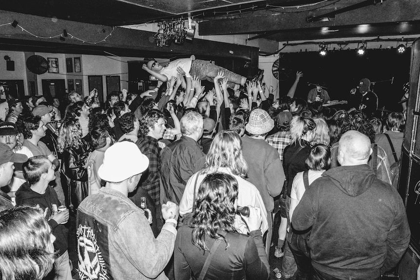 Black and white photo of a chaotic music event with a young man crowdsurfing across a lively crowd