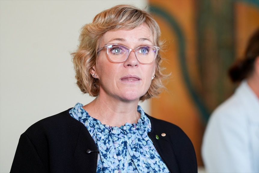 A blonde, formally dressed woman in glasses stands in a room with a large artwork on the wall.