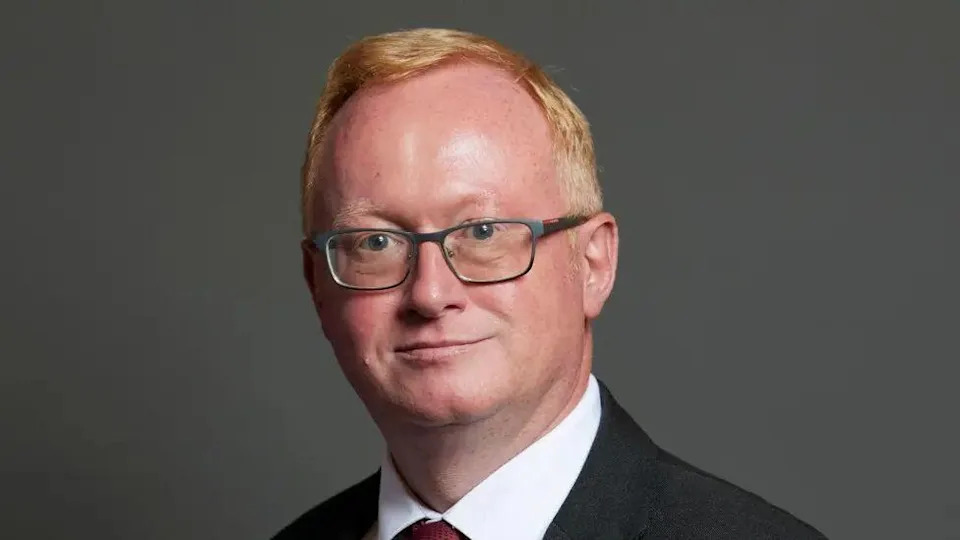 A close-up picture of Luke Akehurst. He has short red hair and is wearing dark-rimmed glasses a white shirt, red tie and black formal jacket.