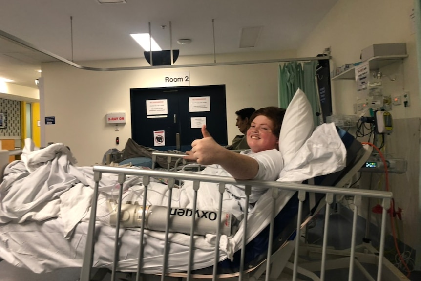 A young boy in a hospital bed, covered in a white sheet gives a thumbs up.