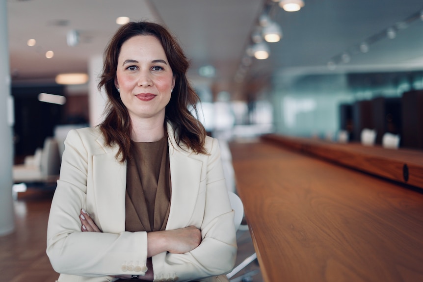 A woman with long brown hair, wearing a cream jacket over a brown top, has her arms folded and smiles to the camera.