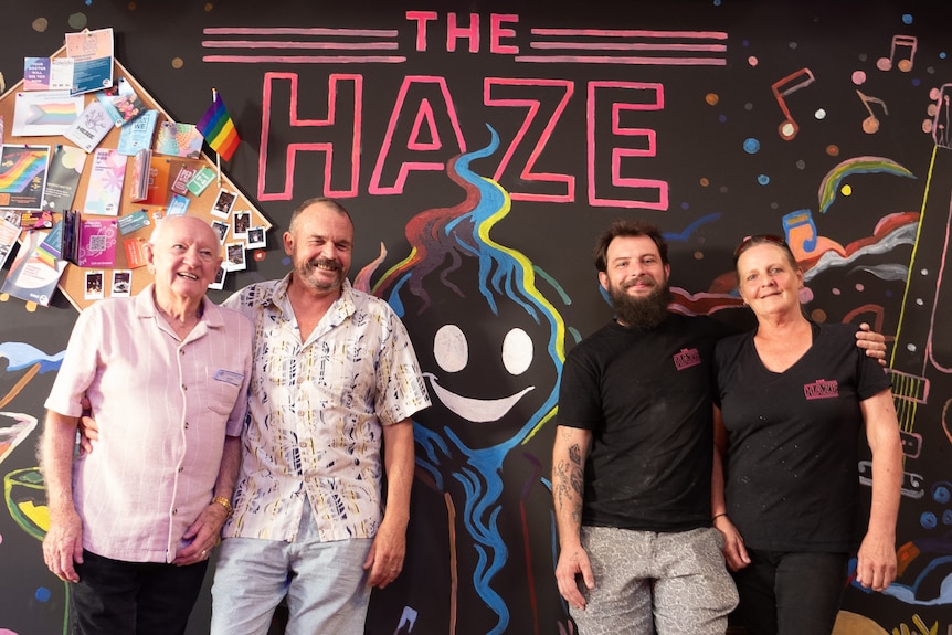 Four people lean against a black wall smiling