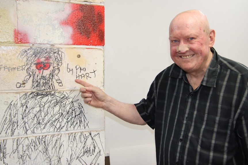 An old man in a shirt points to a drawing of a man on a brick column with the signature 'by Pro Hart'