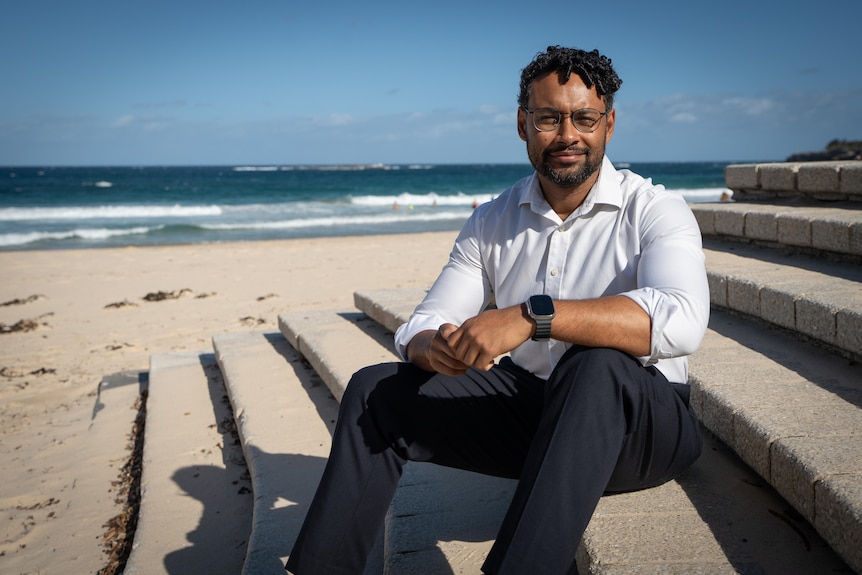 A man sits on steps leading to sand at a beach.