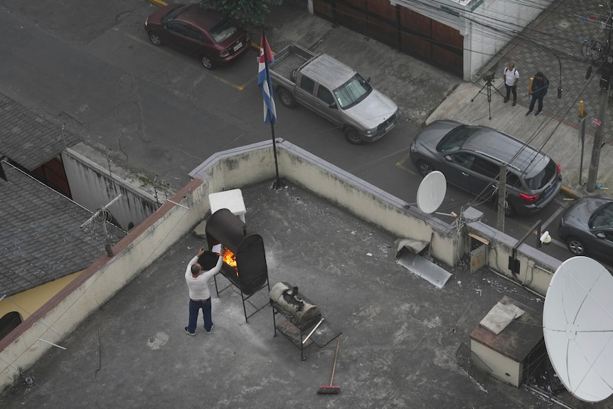 A man in a white top and dark pants putting papers into a burning black barbecue on a rooftop next to a Cuban flag.