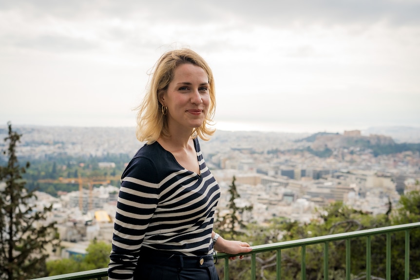 A blonde woman standing with a view of Athens in the background.