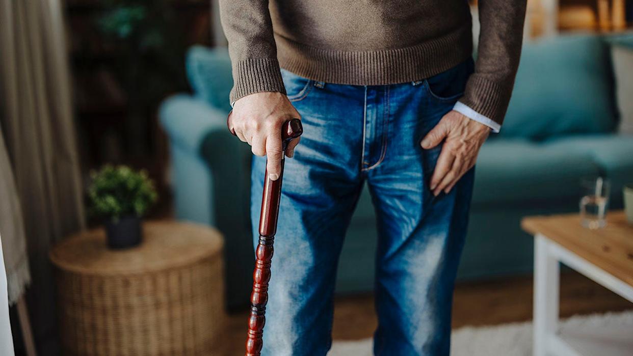 Close-up on elderly person walking with wooden cane, only half their torso and above the knees shown.