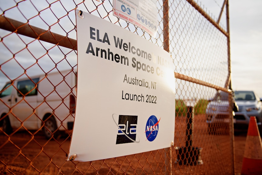 A sign reading 'Arnhem Space Centre' on a wire fence, in front of several cars parked on a patch of red dirt.