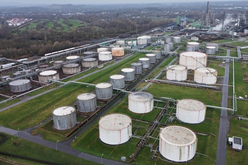 Rows of white oil tanks are seen outdoors in front of a refinery. 