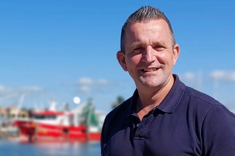 A man smiles as he stands on a wharf with trawlers in the background.