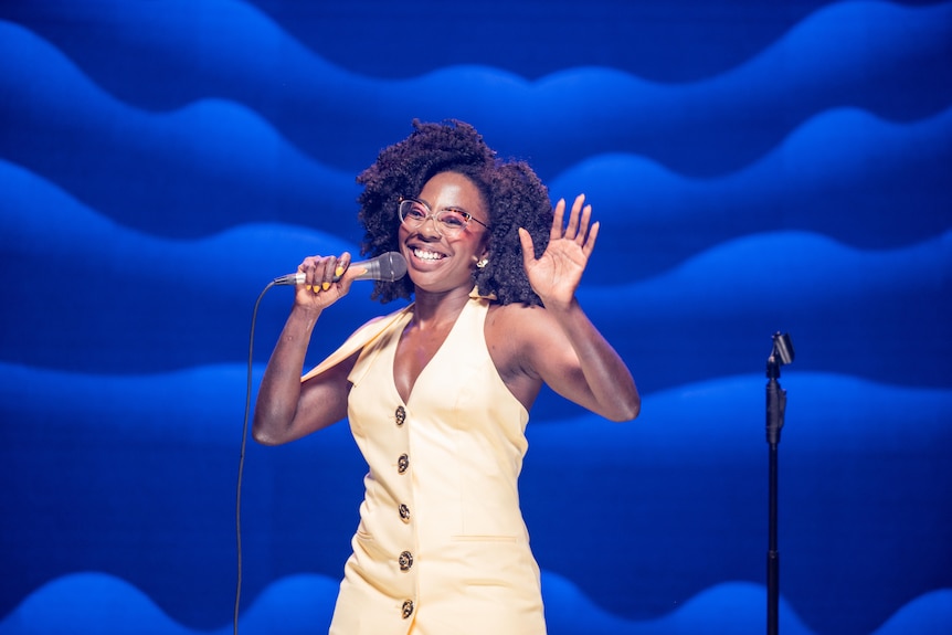 Anisa Nandaula, a Ugandan Australian in her mid-20s, in glasses, smiles brightly on stage, holding a microphone in one hand.