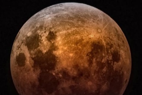Up close image of the moon during an eclipse, showcasing its dark grey speckles against a glowing orange surface