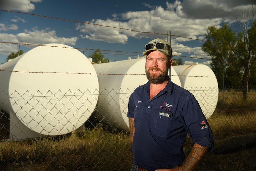 A man in a trucker cap and work shirt stands smiling beside fuel tanks.