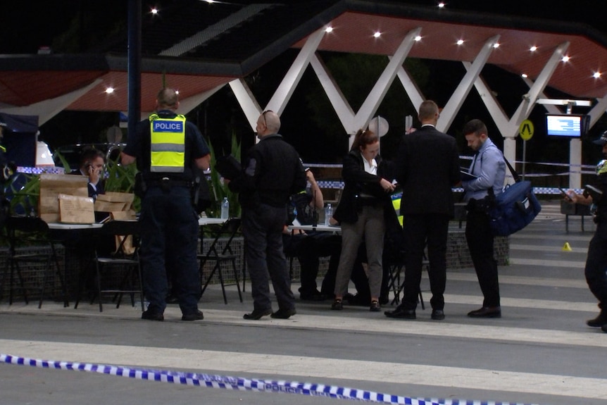 Numerous police officers stand and sit at outdoor dining tables behind police tape at night.