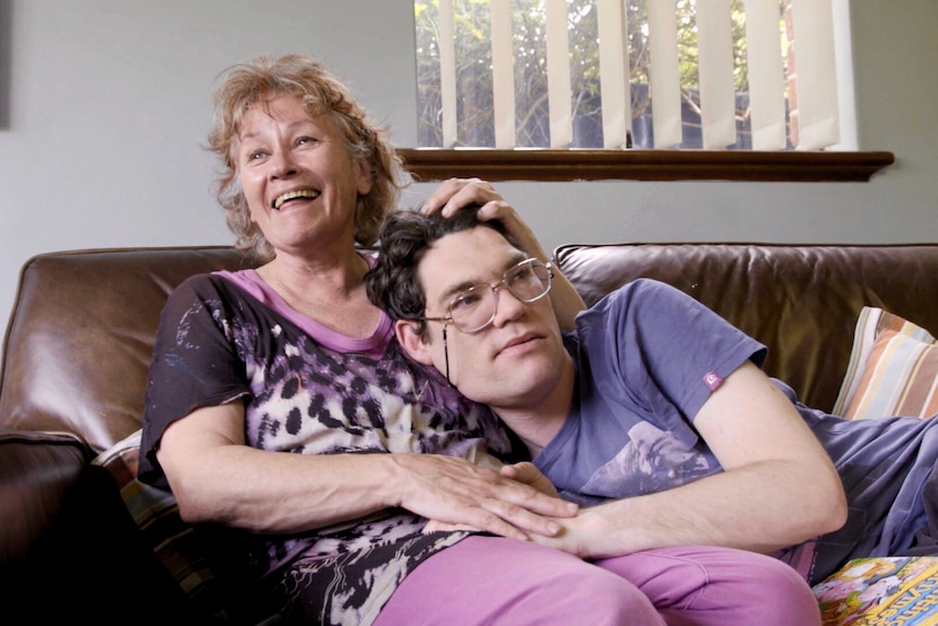 Middle-aged woman wearing pink top and pants smiles looking to distance sitting on a couch. Her adult son rests head on her lap