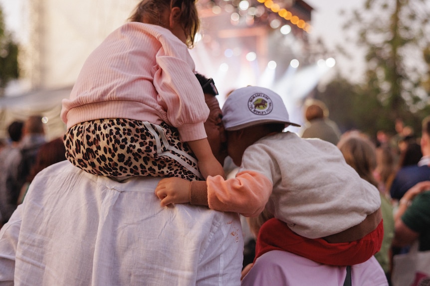 Crowd scene with a person carrying two children on their shoulders while watching an outdoor festival stage.