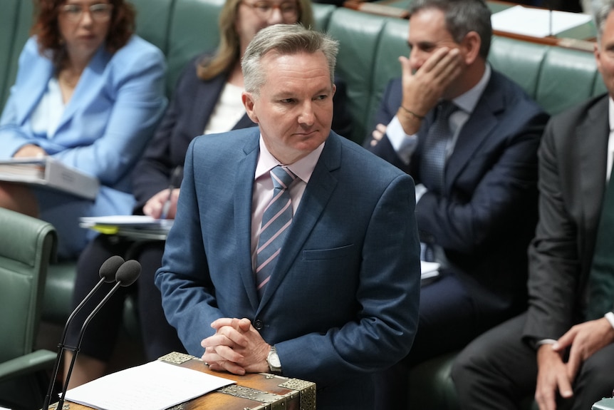 A man in a suit and tie speaking in Parliament, standing in front of a bench of his colleagues.