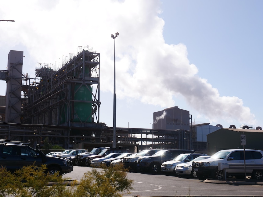 Image of an industrial building on a sunny day. There's a carpark in the foreground and steam billowing from smokestacks.