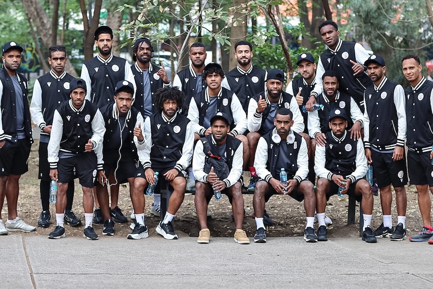 A group of men wearing black and white stadium jackets and grey shorts stand in a group outside posing for a photo