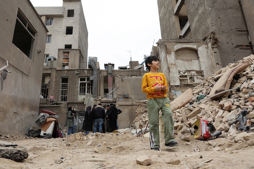 A young boy in a yellow shirt stands amongst rubble