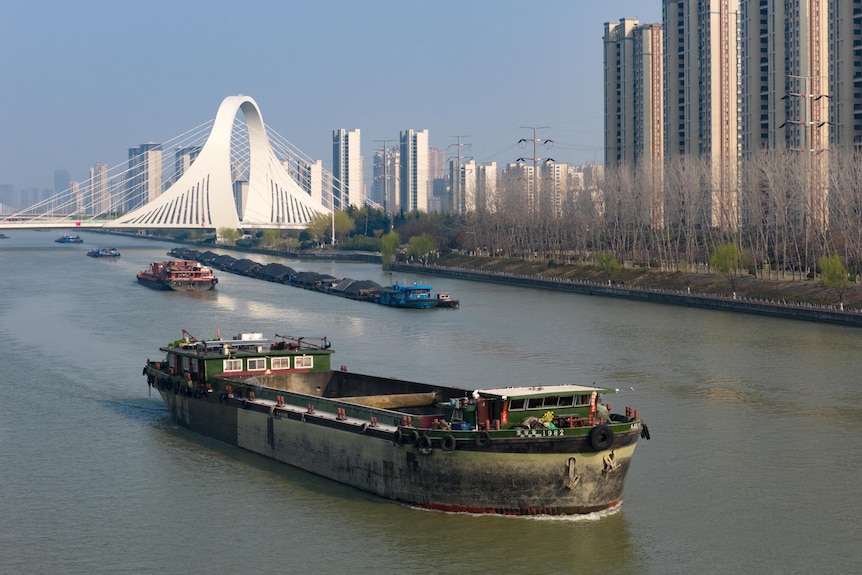 Container ships on a canal in front of a white suspension bridge that has an arcing loop in the middle of its structure.