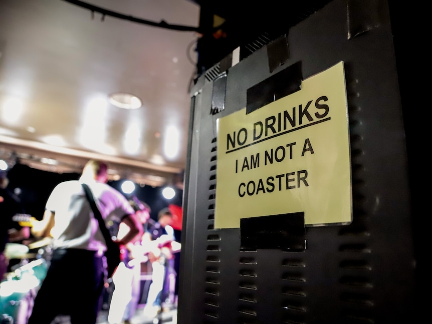 A sign that reads 'No Drinks I am Not a coaster' taped to the back of a stack of speakers behind a live music stage