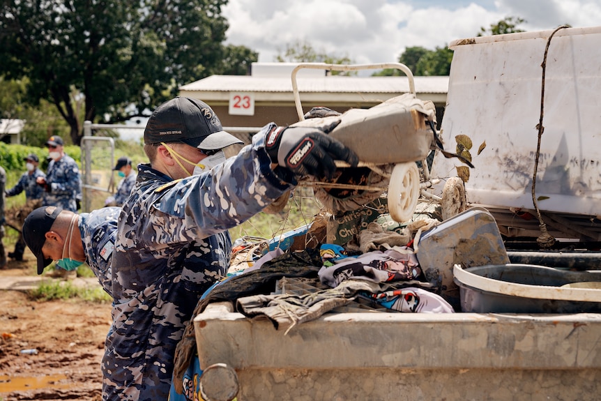 A person wearing a camoflague uniform lifts rubbish into a skip bin.