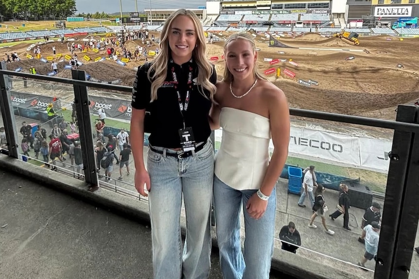 Two younger women standing on a balcony with a motocross track in the background