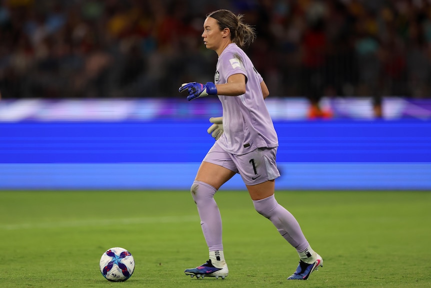 A goalkeeper in grey controls the ball in front of advertising hoardings