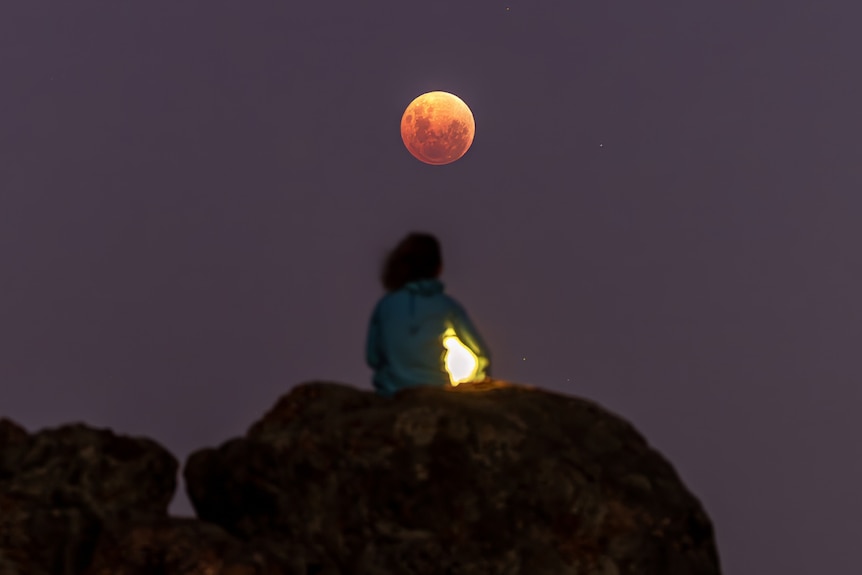 A woman sits on rocks looking at the blood moon.