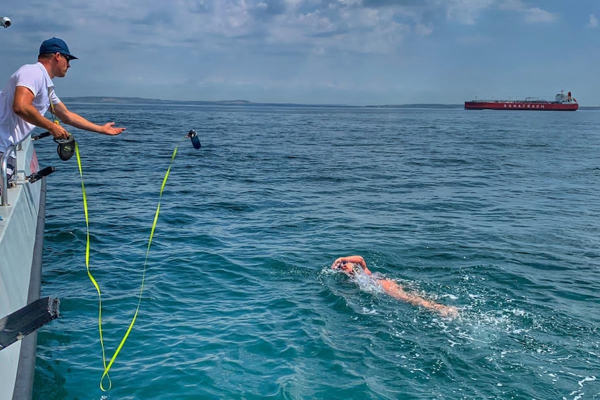 A man stands on a boat over blue water holding a dog lead with food to a swimmer in the water