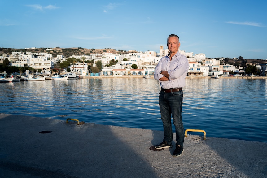 A man stands on a waterfront with a small town behind him.