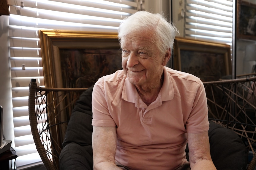 A man in his 80s wears a collared shirt and sits in a wicker chair, smiling and looking to the right