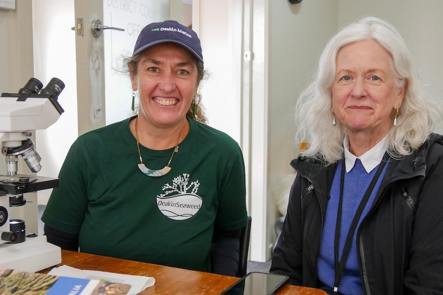 Two women smiling at the camera while sitting next to a microscope. 
