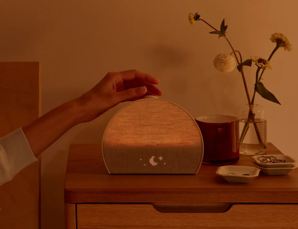 A hand adjusts a minimalist, semi-circular lamp on a bedside table with flowers, a mug, and small dishes