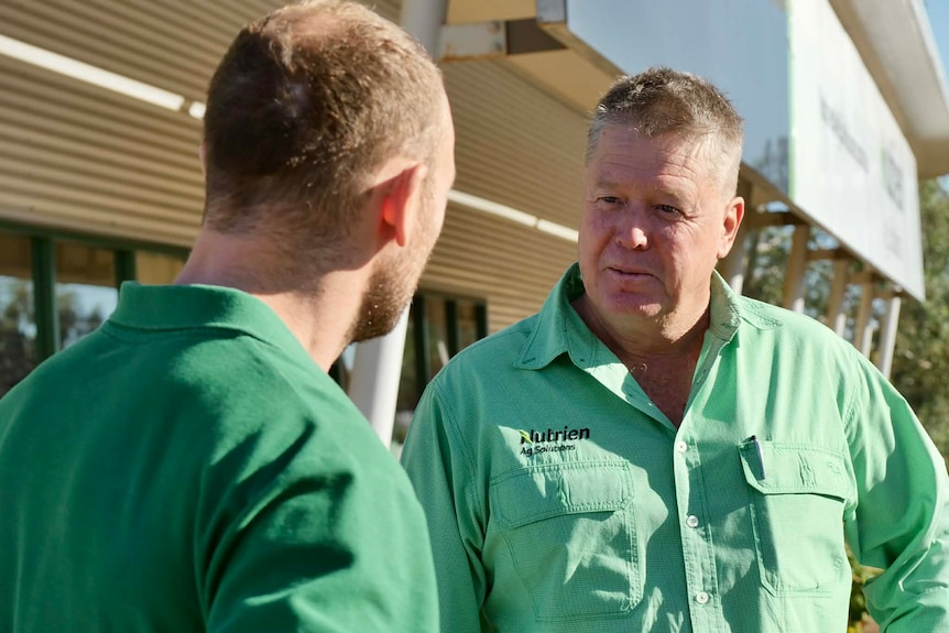 two men in green shirts talking in front of a white building
