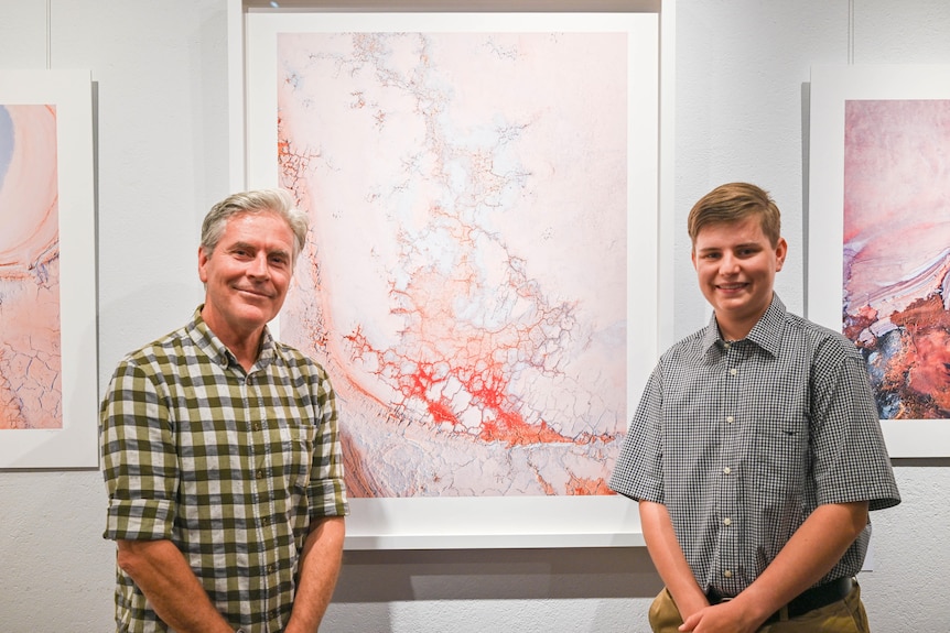 A middle-aged, silver-haired man and a young man with golden-brown hair standing near photographs hanging in a gallery.