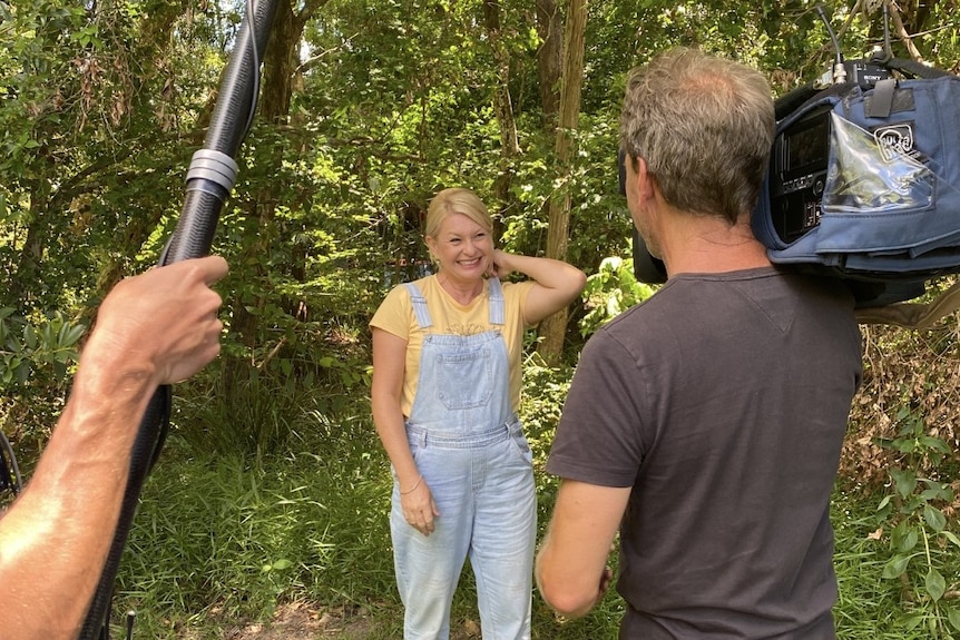 A smiling blonde woman in denim overalls is being filmed by a TV cameraman with a boom microphone held above.