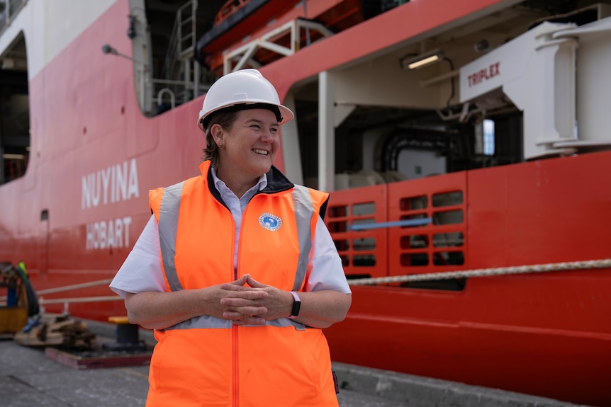 A lady in a high-vis vest and a white hard hat stands and smiles in front of a large red and white ship.