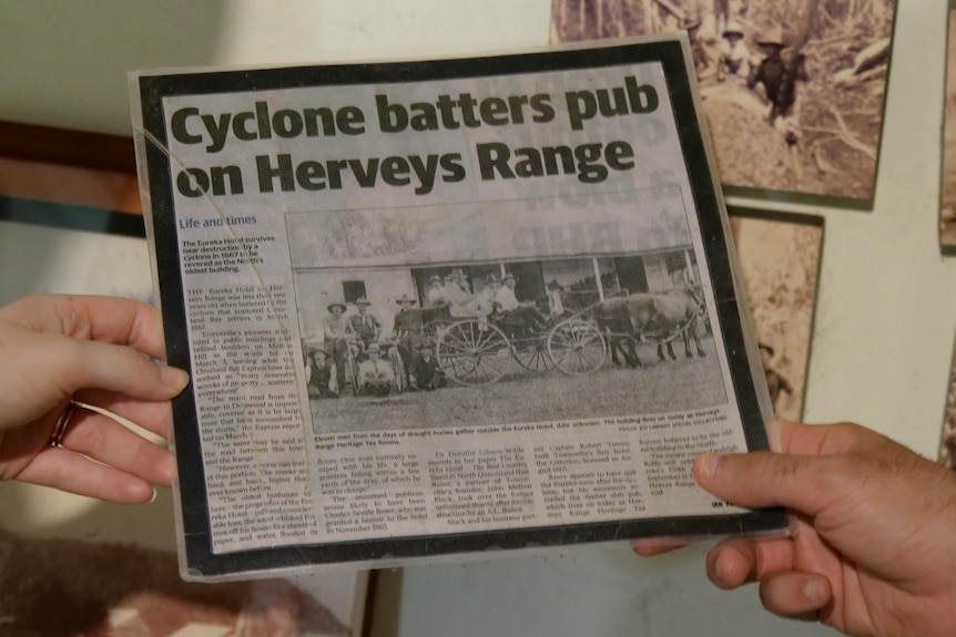 Two hands holding a newspaper clipping that says "cyclone batters pub on Herveys Range".