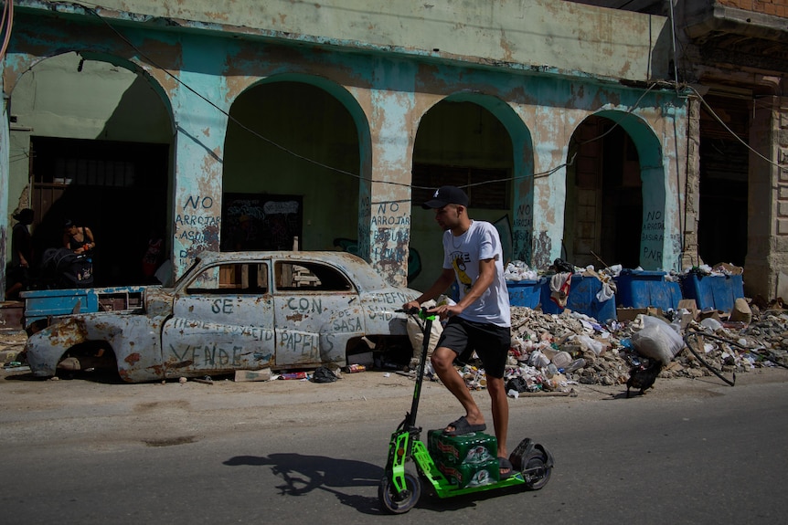 A Cuban man riding a green scooter on a road next to a rusted classic car wreckage without wheels and piles of rubbish.