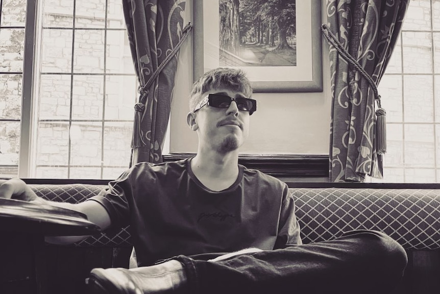 A black and white photo of a young man in dark glasses sitting in a pub.